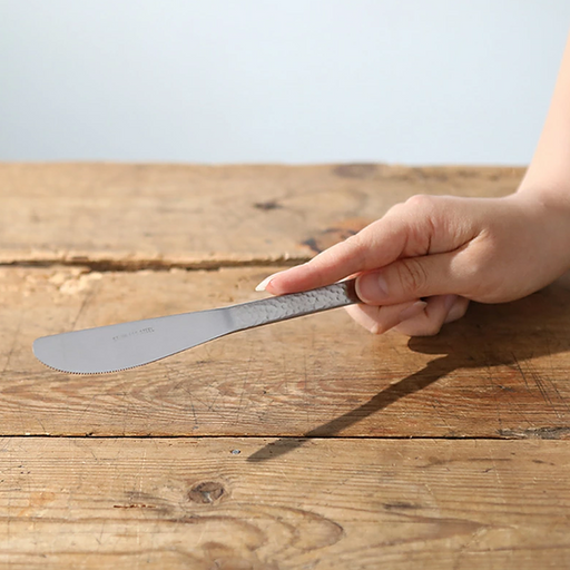 Nagomi Hammered Style stainless steel table knife with tsuchime textured handle and fine serrated edge, held in a hand above a rustic wooden table