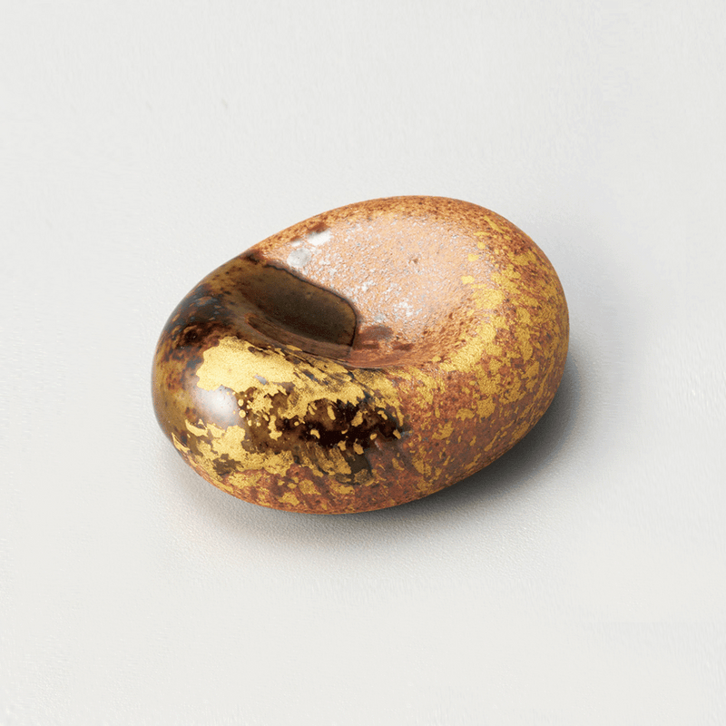 An Oribe Kin-sai chopstick rest, showing its organic bean shape, dark glaze with a touch of green, and patches of gold leaf on a plain background.