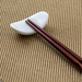 A white ceramic chopstick rest with a swirling pattern, shown holding a pair of dark brown wooden chopsticks, placed on a textured brown fabric surface.