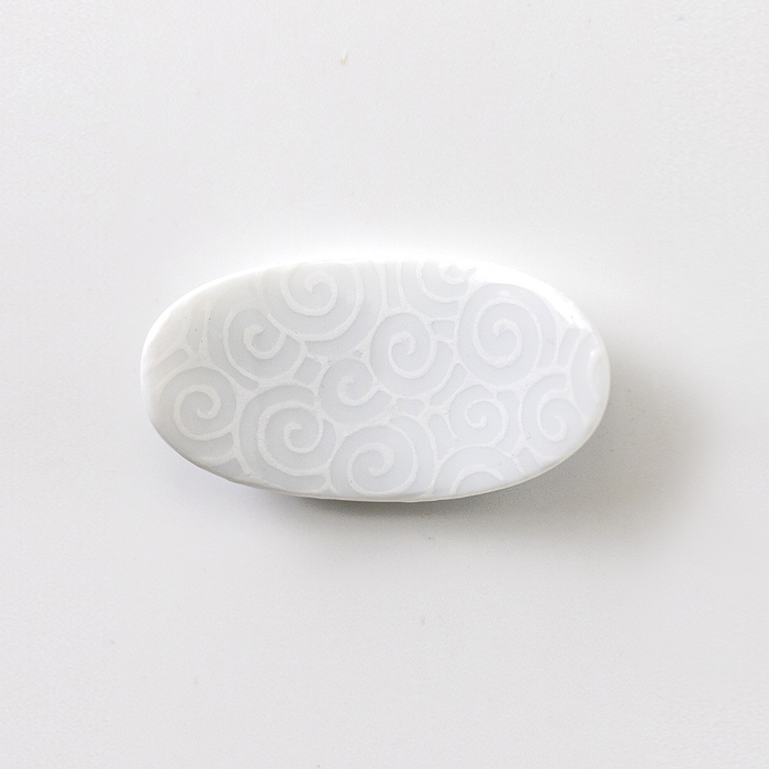 An oval white ceramic chopstick rest with an intricate pattern of raised swirls and curls, creating a textured surface on a plain background.
