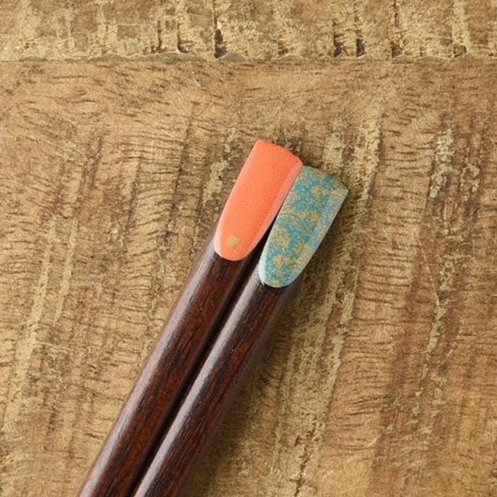 A single pair of wooden chopsticks resting on a wooden table. The coral tip is embellished with intricate gold details, and the turquoise tip displays a delicate gold-flecked leaf pattern.