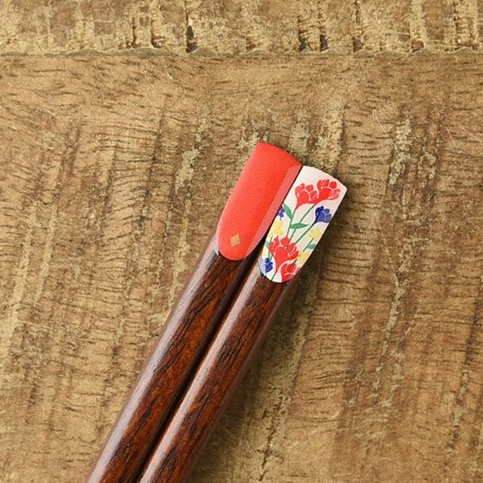 Close-up of wooden chopsticks featuring a coral lacquer tip and a white floral-patterned tip adorned with vibrant red, blue, and yellow flowers.