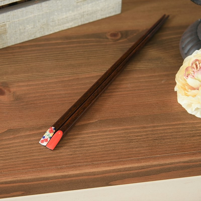 A pair of wooden chopsticks with coral-coloured and floral-patterned tips resting on a wooden table, showcasing intricate red and blue flower designs.