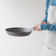 Side view of the River Light Kiwame carbon steel frying pan being held by a person in an apron, showing its depth and wooden handle.