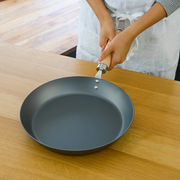 A person wiping the inside of a River Light Kiwame 28cm carbon steel steak pan with a cloth while it rests on a wooden table.