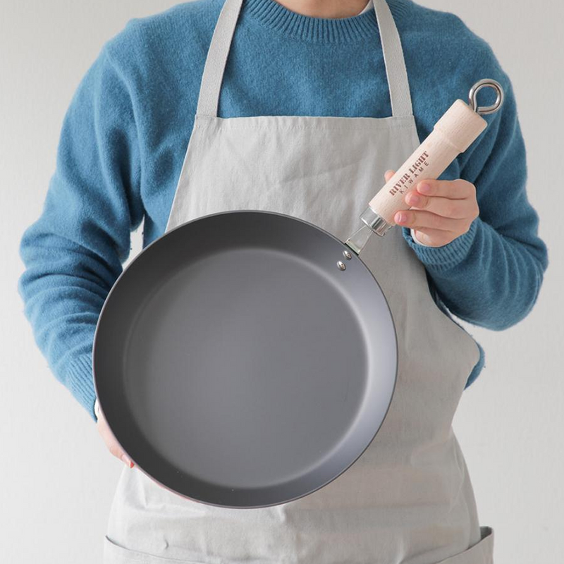 A person in an apron holds the River Light Kiwame 28cm steak pan, showing its large, dark grey carbon steel cooking surface.