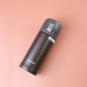 A dark cocoa Zojirushi SM-KB36 vacuum insulated flask with a one-touch open lid lying diagonally on a soft peach background.