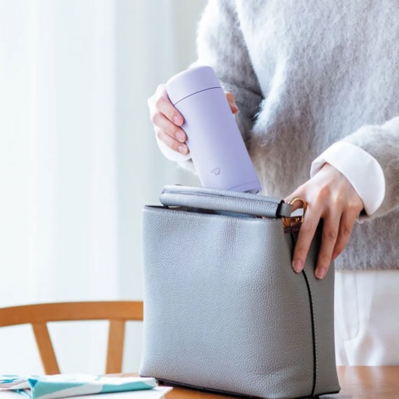 Person placing a Zojirushi Silm Vacuum Insulated Flask - 250ml (SM-MS25-VM) - Purple into a gray bag on a table.