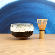 A Japanese Satsuma Tenmoku matcha bowl with a textured white glaze dripping over a matte black base, shown against a plain grey background. Made in Japan