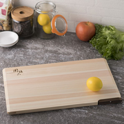 A Seki Magoroku Self-Standing Hinoki Cypress Wood Cutting Board placed flat on a dark gray stone countertop, surrounded by kitchen items like a jar of lemons, an apple, and lettuce. The light, natural-toned Hinoki cypress wood features a smooth surface with a dark walnut lever on one edge. A yellow lemon rests on the board, highlighting its use for food preparation.