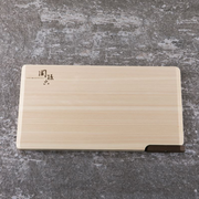 A Seki Magoroku Self-Standing Hinoki Cypress Wood Cutting Board lying flat on a dark gray stone countertop. The light-colored Hinoki cypress wood features a smooth, natural grain with subtle Japanese characters etched on the surface. A dark walnut lever is visible on one edge, designed for self-standing storage, adding a minimalist and elegant contrast.