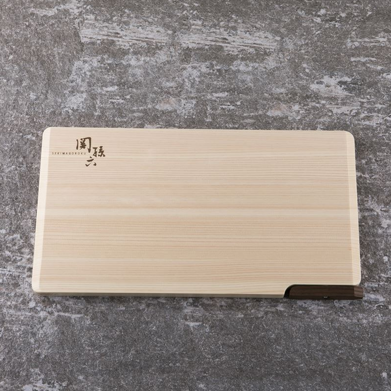 A Seki Magoroku Self-Standing Hinoki Cypress Wood Cutting Board lying flat on a dark gray stone countertop. The light-colored Hinoki cypress wood features a smooth, natural grain with subtle Japanese characters etched on the surface. A dark walnut lever is visible on one edge, designed for self-standing storage, adding a minimalist and elegant contrast.