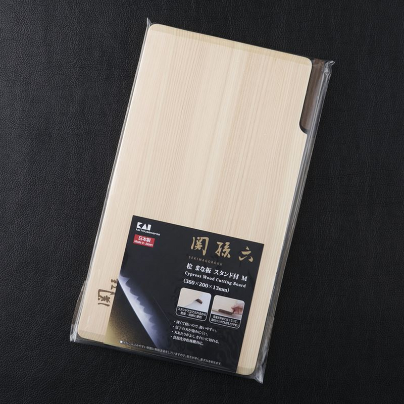 A packaged Seki Magoroku Self-Standing Hinoki Cypress Wood Cutting Board, made in Japan, shown in clear plastic wrap on a black leather background. The board is light-colored with a natural wood grain, featuring a dark walnut lever for self-standing. The packaging includes Japanese text and the brand logo, KAI, with dimensions listed as 360mm x 200mm x 13mm.