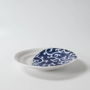 Side view of a white ceramic plate with a bold indigo Karakusa swirl design covering one side and a cat illustration peeking out from the pattern.

