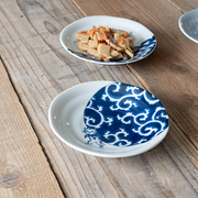 White ceramic plate featuring a dark indigo Karakusa pattern and a small cat illustration near the base, placed on a rustic wooden surface with another patterned plate in the background.

