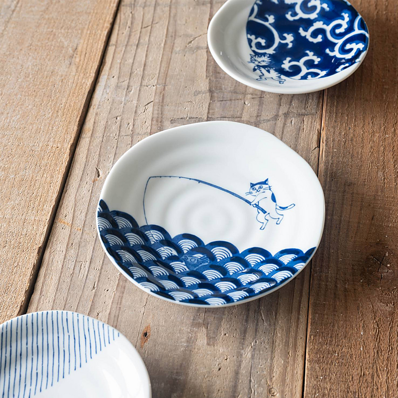Ceramic plate featuring a cat fishing in a seigaiha wave pattern, set on wooden table.


