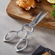 Shimomura Pro-Grade all stainless steel kitchen scissors with serrated bottle opener grip, shown open on a wooden cutting board alongside grilled chicken thigh and lemon.
