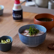 A person holding a blue and white Tokusa-patterned Japanese bowl filled with ramen noodles, using chopsticks to lift noodles, with additional Japanese dishes visible on the table.