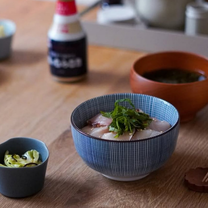 A person holding a blue and white Tokusa-patterned Japanese bowl filled with ramen noodles, using chopsticks to lift noodles, with additional Japanese dishes visible on the table.