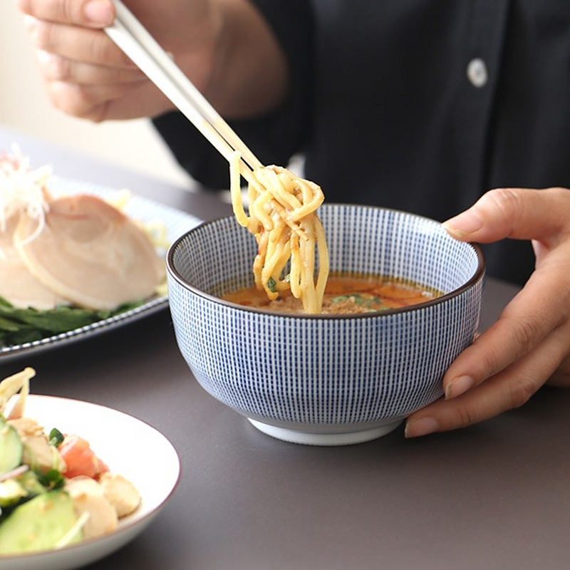 A blue and white Tokusa-patterned Japanese bowl containing sashimi topped with green herbs and sesame seeds, placed on a wooden table with a small dish of pickled vegetables and a bottle of soy sauce in the background.