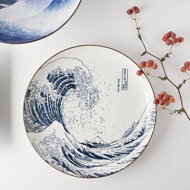 A 25cm porcelain dinner plate with an iconic depiction of The Great Wave off Kanagawa, featuring indigo blue on a white background, with fine waves curling over Mount Fuji in the background. The plate has a smooth glazed surface with a subtly elevated rim.