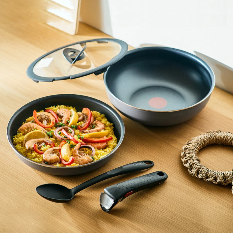 A T-fal Ingenio frying pan and wok on a wooden table with paella, a glass lid, detachable handle, and cooking spoon.

