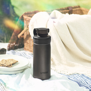 A steel grey Tiger MTA-T050 vacuum insulated carbonated bottle with a black screw-top lid and integrated loop handle, standing on a striped picnic blanket with a wicker basket in the background.