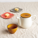 A table setting with a mustard-colored teacup, a matching mustard-lidded teapot, and colorful plates holding small pieces of cake, set on a beige cloth background.