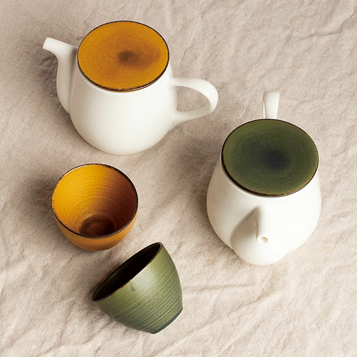 A close-up of a green teacup leaning on its side next to a mustard teacup, with matching lidded teapots in the background, all set on a beige fabric.