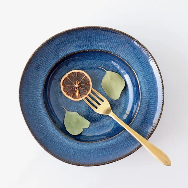 A close-up view of a Tojiki Tonya Orlo Series Pasta Plate, 24cm, showcasing a lemon slice and two green leaves on the deep blue ceramic surface with a golden fork placed on the plate.