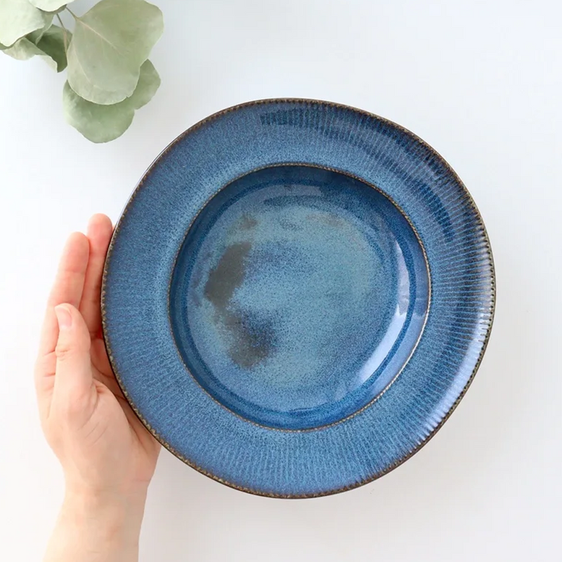 A hand holding a Tojiki Tonya Orlo Series Pasta Plate, 24cm, highlighting the deep blue ceramic surface with a textured rim, set against a white background with green leaves in the corner.