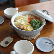 An inviting scene displaying a meal served in the Tojiki Tonya Tstyle Donabe Japanese clay pot. The open pot reveals a steaming dish with vegetables, proteins, and garnishes. A white plate, chopsticks, and a white mug accompany the presentation on a wooden table.