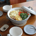 An inviting scene displaying a meal served in the Tojiki Tonya Tstyle Donabe Japanese clay pot. The open pot reveals a steaming dish with vegetables, proteins, and garnishes. A white plate, chopsticks, and a white mug accompany the presentation on a wooden table.