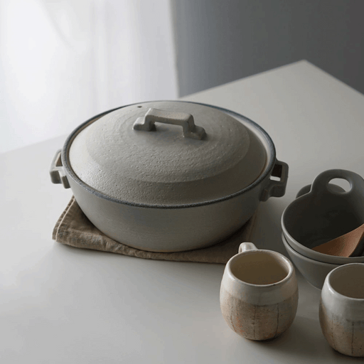 Isolated front view of the Tojiki Tonya Tstyle Donabe Japanese clay pot on a light backdrop. The pot's silver-grey texture and its circular form are evident, with the pot's handles and lid handle in full view.