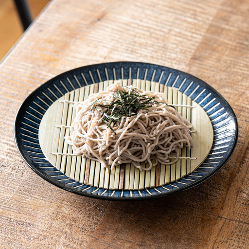 A large 25cm Toki Takane porcelain plate featuring a traditional indigo blue 'Tokusa' stripe pattern, displayed with a round bamboo-style zaru draining mat for serving cold soba noodles.