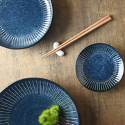 A tablescape featuring various sizes of the Tokusa Indigo plates arranged on a dark wood surface with chopsticks.

