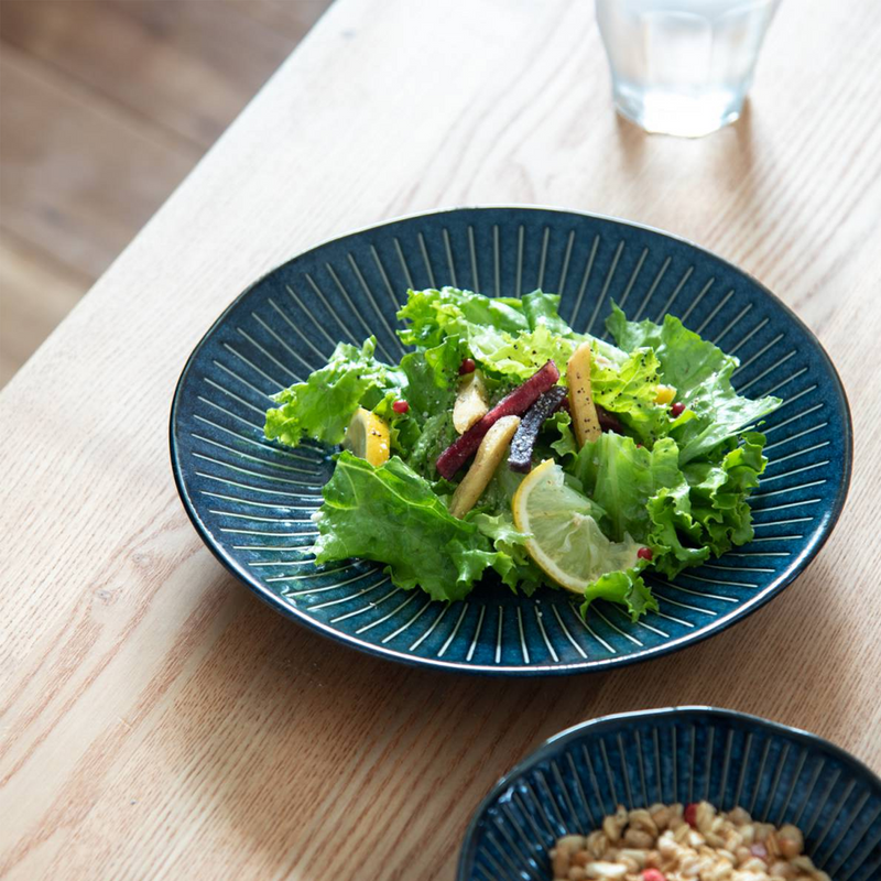 A fresh green salad served on the Tokusa Indigo Dinner Plate (23cm), which is sitting on a light wood dining table.

