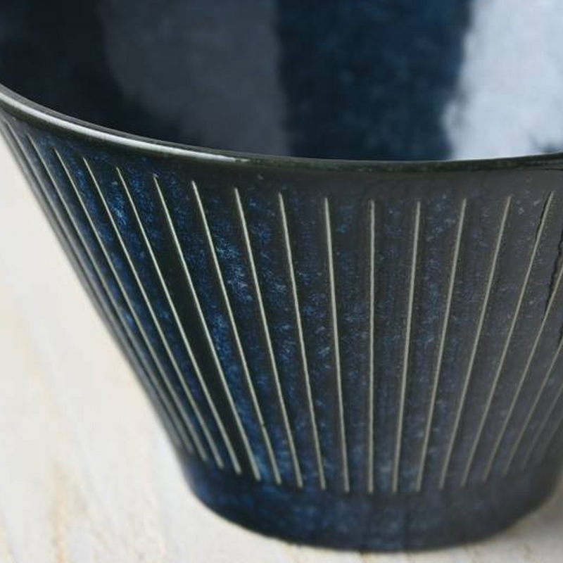 A close-up macro shot of the Tokusa Indigo Salad Bowl, highlighting the detailed vertical stripes and the rich, speckled indigo glaze.

