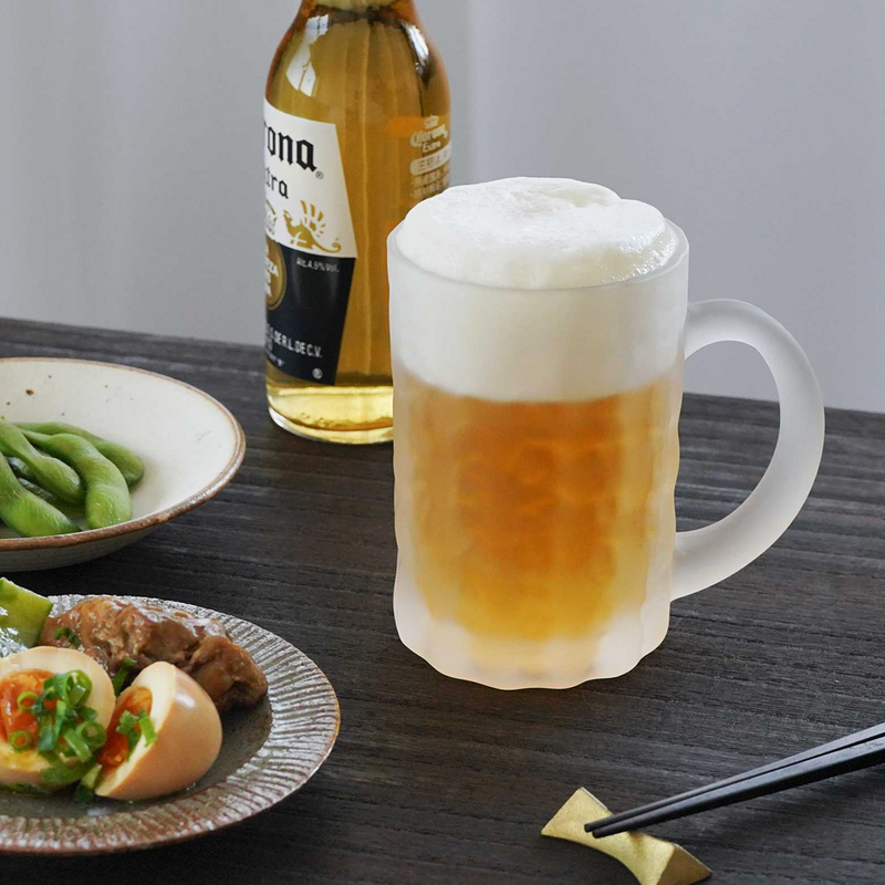 Toyo Sasaki Frosted Beer Mug in Use with Beer and Food – A frosted beer mug filled with golden beer and a thick foamy head, placed beside Japanese appetizers, with a beer bottle in the background.


