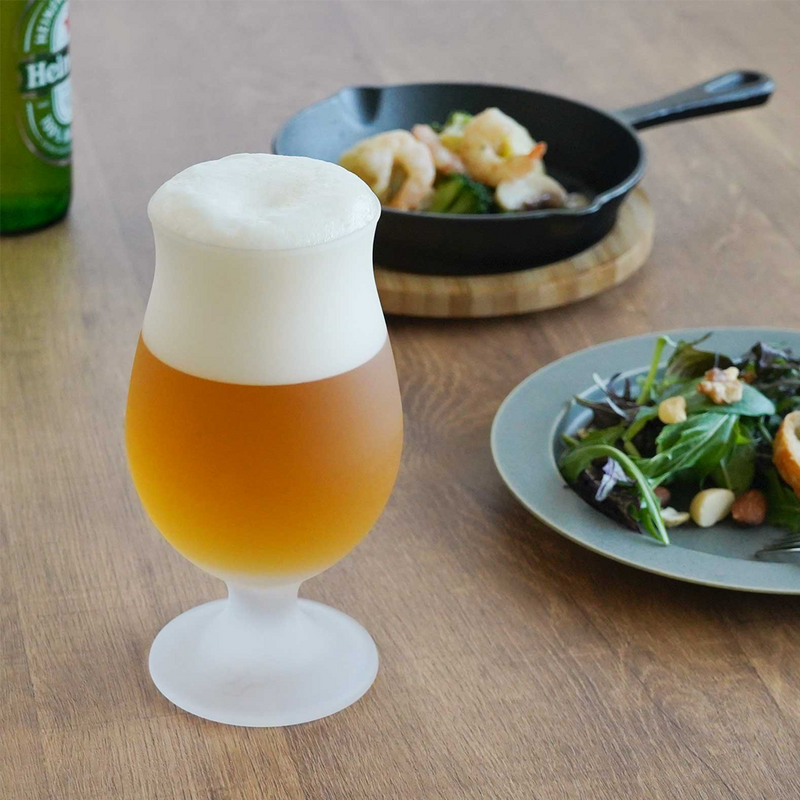 Frosted tulip beer glass filled with golden beer and topped with foam, placed on a wooden table with a meal in the background.

