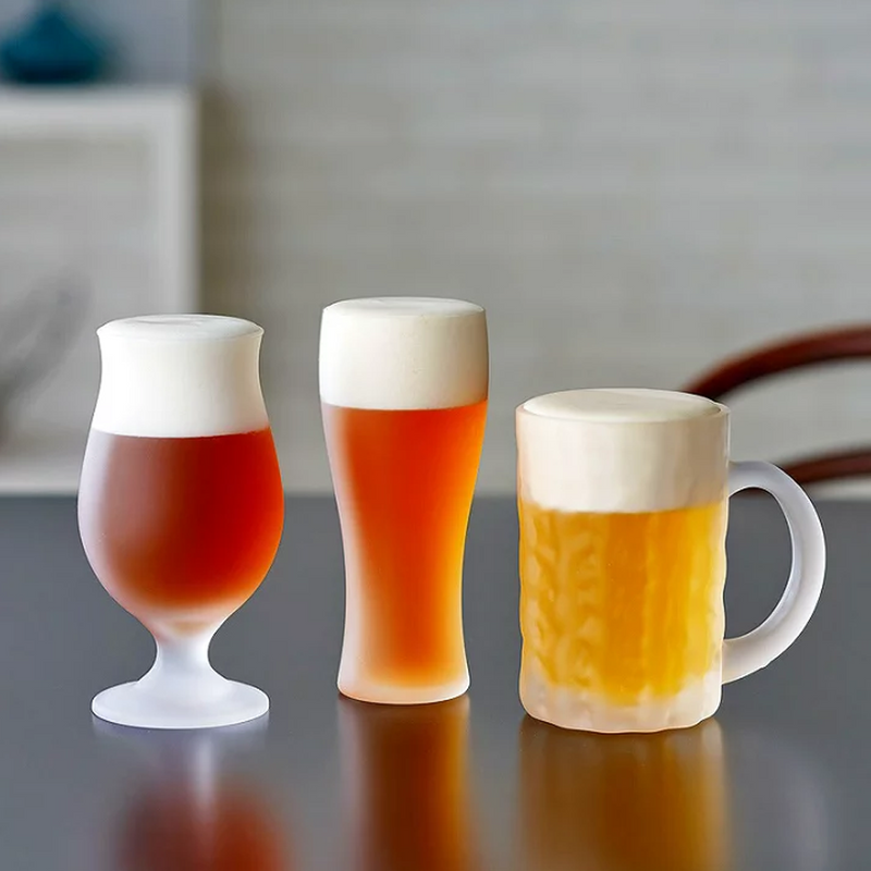 Three frosted beer glasses – tulip, pilsner, and mug – filled with beer and topped with thick foam, placed on a reflective surface.
