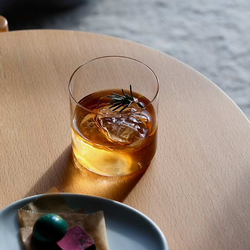 Toyo Sasaki USURAI On The Rock Glass with an ultra-thin rim, filled with whisky, a large ice cube, and a rosemary sprig, on a round wooden table beside a plate of sweets.

