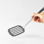 A hand holding the Tsubame Sanjo Stainless Steel Slotted Turner to demonstrate its size and single-piece design against a white background.

