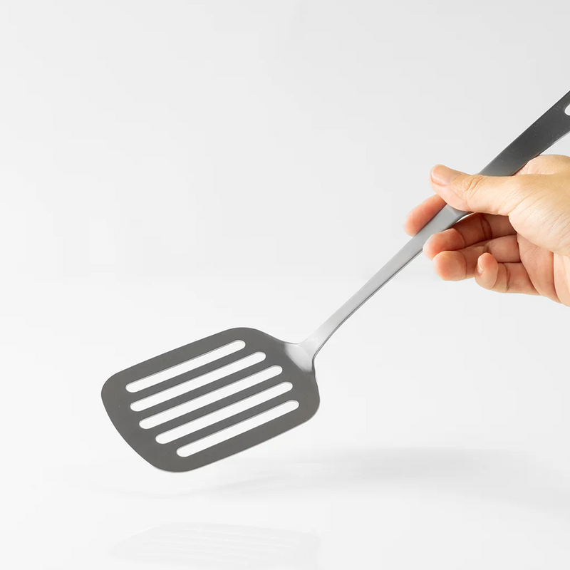 A hand holding the Tsubame Sanjo Stainless Steel Slotted Turner to demonstrate its size and single-piece design against a white background.

