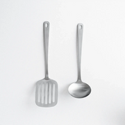 Two matte stainless steel kitchen utensils, a slotted turner and a soup ladle, arranged side-by-side on a white background.
