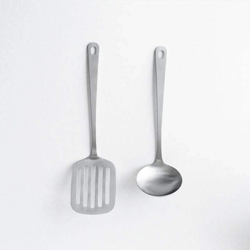 Two matte stainless steel kitchen utensils, a slotted turner and a soup ladle, arranged side-by-side on a white background.