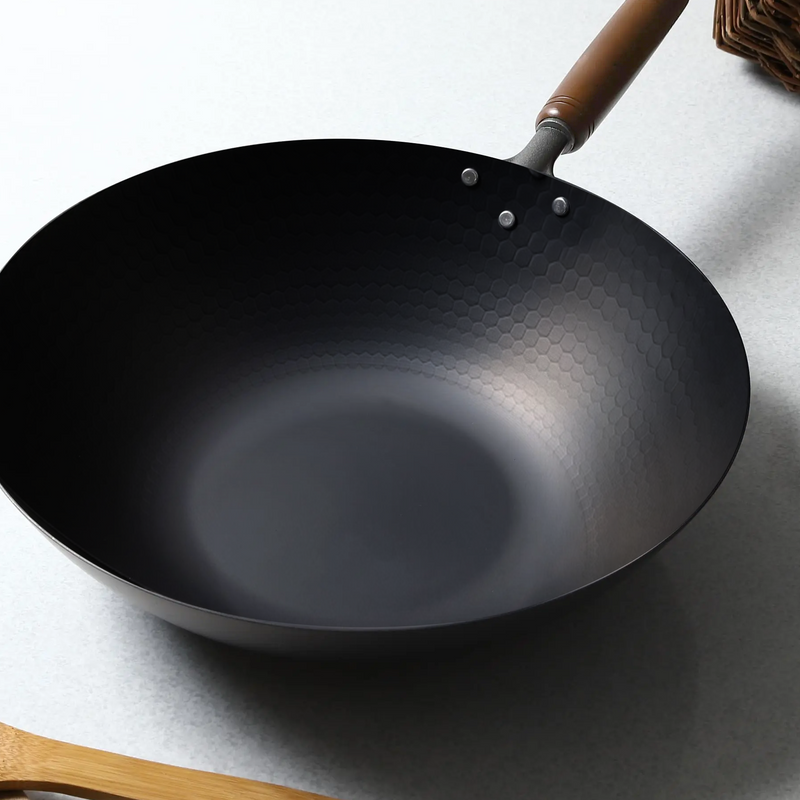 A Tsubame Sanjo hammered style nitrided carbon steel induction wok, 30cm, with a wooden handle, sitting on a light grey surface.
