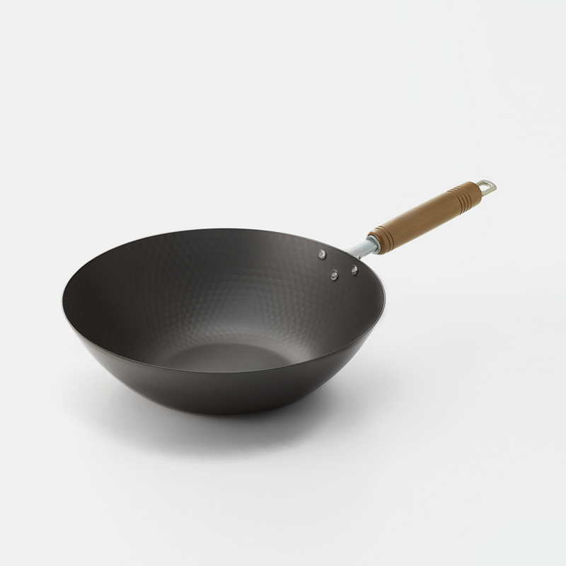 Black wok with a wooden handle on a white surface, with a wooden spoon in the foreground and a woven basket in the background, top-down view.