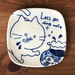 Top view of a square ceramic plate with a hand-drawn indigo blue cat design and the text 'Let’s get along well' surrounded by playful patterns, including a fish and splashes.