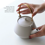A person attaching the brass handle to a white Yamatsu Frustum Japanese teapot, showing the side slot mechanism.